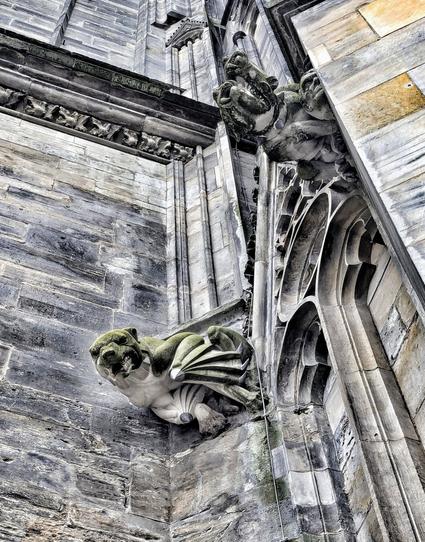 A low-angle, close-up shot captures the intricate architectural details of a Magdeburg Cathedral. The structure is predominantly grey, with variations in tone suggesting age and exposure to the elements.
The focal point is a grotesque gargoyle figure, positioned on the left side of the frame. It is carved from the same grey stone as the building and features a somewhat humanoid face with large eyes and a downturned mouth, combined with bat-like wings that extend downwards. Patches of green moss or lichen are visible on its surface, indicating its age and exposure to moisture.
To the right of the gargoyle, a section of the building's facade shows ornate Gothic-style arched windows or niches, also constructed from grey stone. Above these architectural elements, more detailed stonework and mouldings are visible, running horizontally across the building's surface. The texture of the stone is rough and uneven, with signs of weathering.