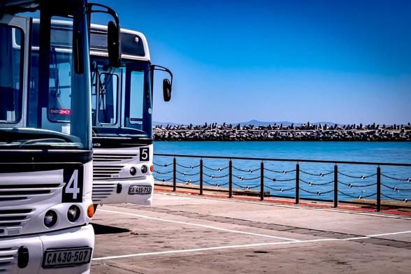 The image shows two parked buses numbered 4 and 5 in a parking area near a body of water. In the background, there is a barrier along the water's edge, and the sky is clear and blue.
