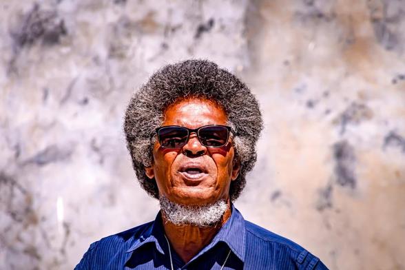 The image shows a person wearing sunglasses, with curly hair and a beard. They are in front of a wall with a textured pattern, wearing a blue shirt.