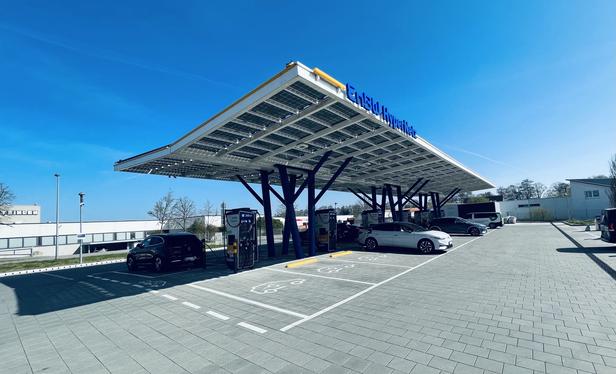 A modern electric vehicle charging station with a solar panel roof is shown. Several parked cars are visible, ready for charging. The sky is clear and blue.