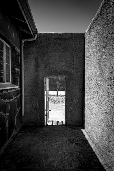 The image shows a black and white photograph of a narrow alley or passage between two buildings. There's an open door leading to a sunlit exterior area. The composition emphasizes strong contrasts between light and shadow, creating a dramatic and moody atmosphere.