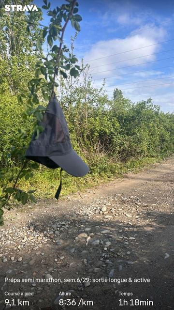 Une casquette grise est accrochée à une ronce à hauteur d'homme. En arrière plan, un chemin de terre bordé de végétation sous un ciel printannier. Des statisiques Strava sont visibles.