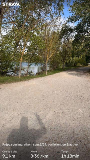 Un chemin de terre sépare les eaux bleu d'un lac et une bande de végétation. Sur le sol, l'ombre du photographe faisant le v de la victoire avec ses doigts. Des statisiques Strava sont visibles.