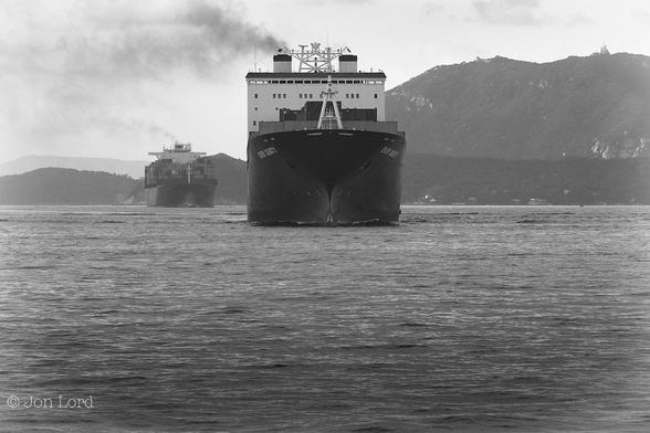 This Is A Black And White Seascape Photo In Landscape Format. Filling The Lower Half Of The Image Is A Body Of Fairly Still Dark Water. In And Above The Centre Is The Bow Of A Large Container Ship, Underway, Heading Exactly Towards The Camera And Perhaps Five Hundred Metres Away. In Common With Many, The Ship Has A Dark Hull And A White Superstructure That Is Towards The Stern. There Are Some Shipping Containers Visible Stacked Above Deck, But Not Any Where Near Fully Loaded. Behind The Bridge Are Two Square Funnels, One Emitting Dark Smoke. About A Kilometre Behind This Ship Is Another Larger Container Ship, Similar To The First Apart From Having A Great Many Containers Stacked Above Deck And Only One Funnel, This To Emitting A Trail Of Dark Smoke.To The Right And In The Background Is Land With Rising Terrain. Above Is A White And Overcast Sky.
The Location Is The East Lamma Channel That Leads From The Deep Waters Of The South China Sea To Hong Kong (2018).