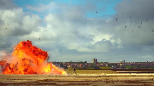 [Image extraite de la série anglaise "Utopia" (Dennis Kelly au scénario), extraite de la saison 1- épisode 2 ("Le manuscrit" ; 2013). Plan géneral présentant à gauche une voiture entrain en feu, en pleine explosion, et au centre une silhouette (d'une femme, autrice de cet acte) marchant d'un pas décidé de gauche à droite.]