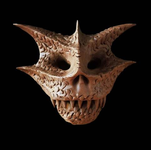 This image shows a sculptural mask with a demonic or monstrous appearance, crafted from brown plasticine. The mask features sharp, pointed protrusions extending from the top and sides, giving it a crown-like or horned appearance. It has hollow eye sockets, a nose cavity, and a prominent mouth full of pointed teeth arranged in a menacing grin. The surface is textured with scale-like or organic patterns carved throughout. The mask is photographed against a solid black background, creating a dramatic contrast that highlights its detailed features and three-dimensional form. This type of mask appears to be inspired by fantasy, horror, or mythological themes, possibly designed for decorative, costume, or artistic purposes.