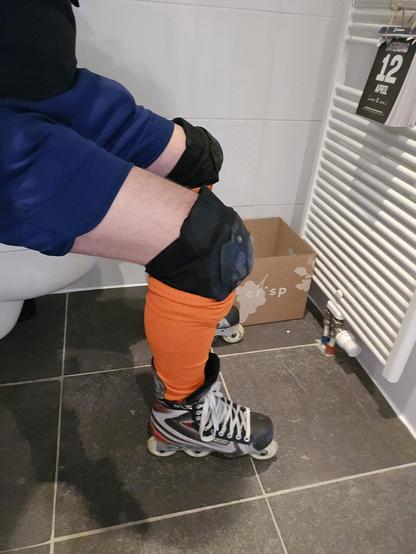 me sitting on my toilet while wearing inline skates. My body is visible from the skates on the floor to my waist. wearing my shorts, knee pads, and bright orange socks. My knee is bend at a comfortable 90 degree angle. Visible other than me, there is a tear-off calendar hanging on a vertical radiator, and a box for the teared off papers.