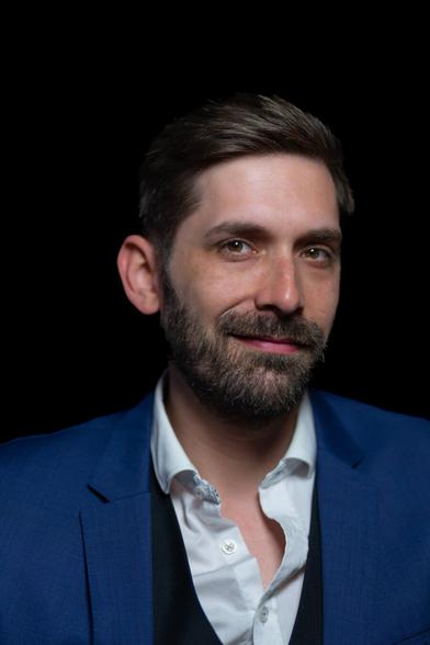 A close-up of a person with short, styled hair, wearing a white shirt, a vest, and a blue suit jacket, offering a slight smile while looking at the camera. The shot is cropped around their upper torso, and the black background frames them in a softly lit setting.