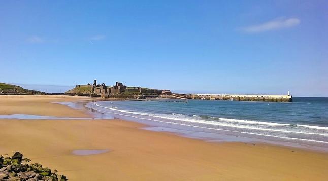 A beach of golden sand, the sea and sky are both bright blue. In the distance, a ruined ancient castle sits on a small island.
