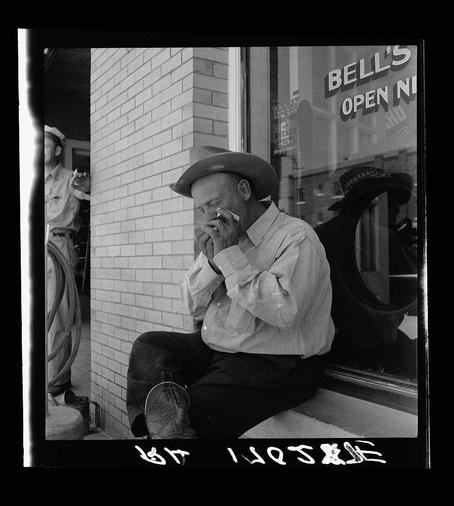 A black and white photograph depicting an older man with a mustache wearing cowboy boots, sitting on the window ledge of "Bell's" establishment. He is smoking a cigarette thoughtfully.

The image captures a candid moment in what appears to be Van Horn, Texas, highlighting cultural or regional elements such as the distinctive boot style worn by this individual.