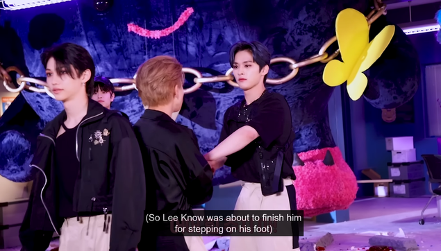 On a MV set, Lee Know has taken hold of Han's wrists and is looking at him sternly. Caption: (So Lee Know was about to finish him for stepping on his foot)