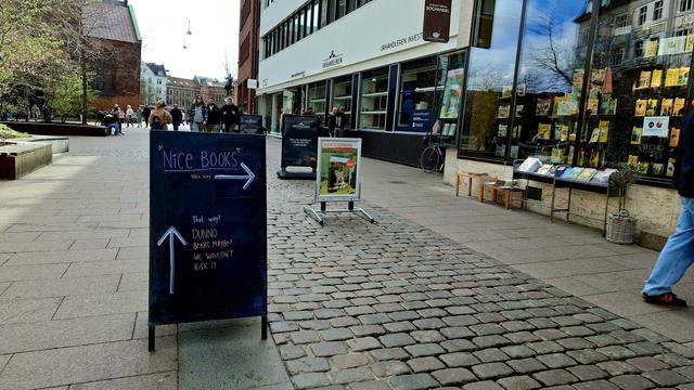 Chalkboard on the cathedral square, mid-city Aarhus. Cobblestones, benches, bikes and pedestrians seen beyond. On the chalkboard, hand-drawn with pretty decorations, in big writing, the headline "Nice Books". Underneath in small writing: "This Way" and a big arrow pointing right. To the right, the window display and pavement book trolleys of Kristian F Møller bookshop.  Below the "Nice books" arrow pointing right: another arrow. This one points straight ahead to the street and square beyond. Written beside it: "That way? Dunno. Bears. Maybe? We wouldn't risk it." To the right, we see the back leg of someone in blue jeans who has just stepped into the book shop, wisely avoiding the bears.
