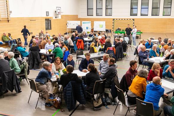 Fotografie von der Bühne in das Publikum. An verteilten Tischinseln sitzen Menschen. Auf den Tischen liegen große weiße Plakate, es stehen Blumen und Getränke drauf. Der Fußboden ist mit bunten Linien überzogen. Im Hintergrund stehen ein Tor, Metaplanwände und ein Tisch voller Getränke.