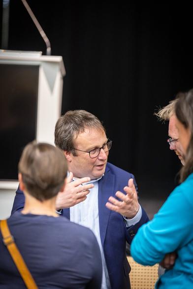 Fotografie von Oliver Krischer oi Gespräch mit drei Personen, die um ihn herum stehen. Im Hintergrund sieht man eine Bühne mit einem Rednerpult.