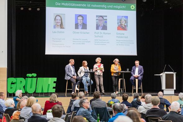 Fotografie einer Bühne mit 5 Personen und einer Leinwand im Hintergrund. Links auf der Bühne stehen grüne Buchstaben, die das Wort "GRÜNE" bilden. Auf der Leinwand sind Fotos von 4 Personen auf der Bühne. Links Lea Dohm, Psychologin und Therapeutin und Mitbegründerin von Psychologists For Future. Dann Oliver Krischer, Minister für Umwelt, Naturschutz und Verkehr des Landes NRW, Prof. Dr. Roman Suthold, ADAC Nordrhein e.V. Verkehrsreferent und Hochschullehrer und Simone Holderried, Bürgermeisterkandidatin und Fraktionsvorsitzende der Brühler Grünen. Über den Fotos steht weiß auf grün "Die Mobilitätswende ist notwendig und möglich!" und rechts davon die gelbe Sonnenblume der Grünen.

Vor der Bühne sitzen viele Personen. Auf den Tischen stehen Blumen und Getränke, einige haben ihre Jacken über die Stuhllehnen gehängt. Rechts auf der Bühne steht ein weißes Rednerpult.