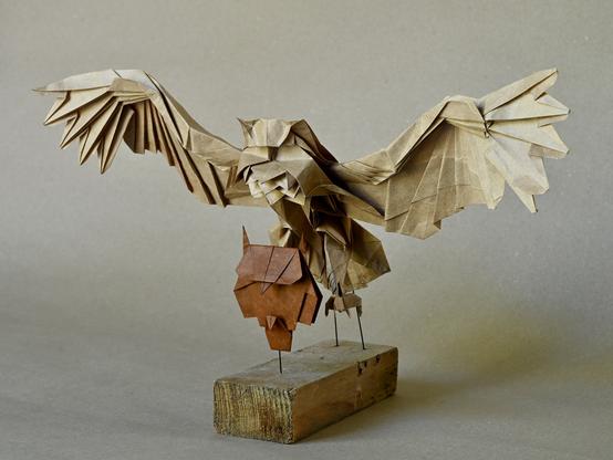 Two origami owls, one modeled in a very detailed way, with spread wings and a feathered body, with fangs ready to grip. The second, smaller one in front of the first is simple and abstract. Both are positioned on a wooden stand.