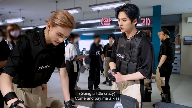 On the MV set, Hyunjin is looking down at some things on a desk. Lee Know is standing nearby and looking at him. Caption: (Going a little crazy) Come and pay me a kiss