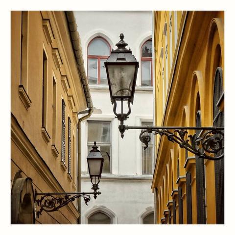 This is a square format colour image of ornate street lamps reaching out across a narrow street. The two lamps are attached to buildings which are painted a warm yellow, one to each side of the frame. A plain stone front building is visible at the end of the street. Taken in Budapest, Hungary.