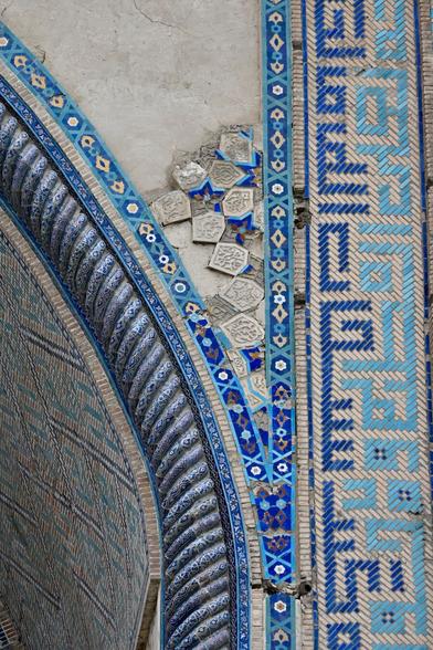 Detail view of the portal of a mosque, decorated with glazed tiles. Some of the tiles have fallen off, leaving parts of the wall exposed.