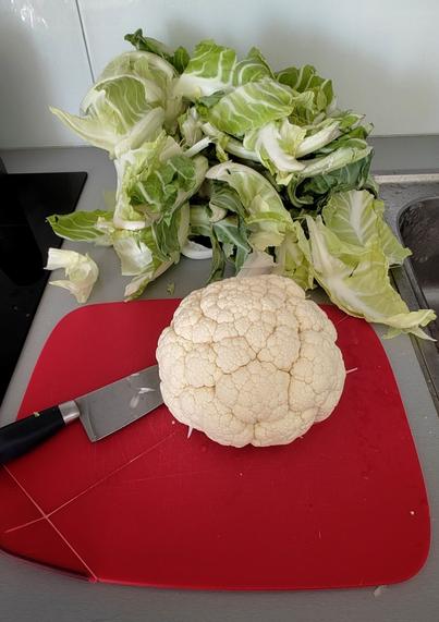 Un choux-fleur épluché sur une planche rouge, en arrière plan un gros tas de feuilles de choux-fleur.