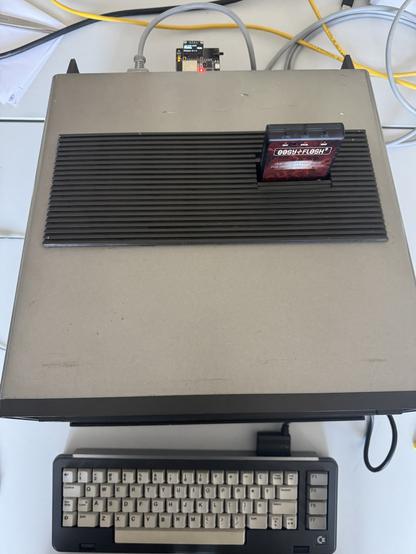 An overhead view of a vintage computer with a cartridge inserted on its top surface. A keyboard is positioned below the computer. Several cables are connected, suggesting a setup for use or testing.