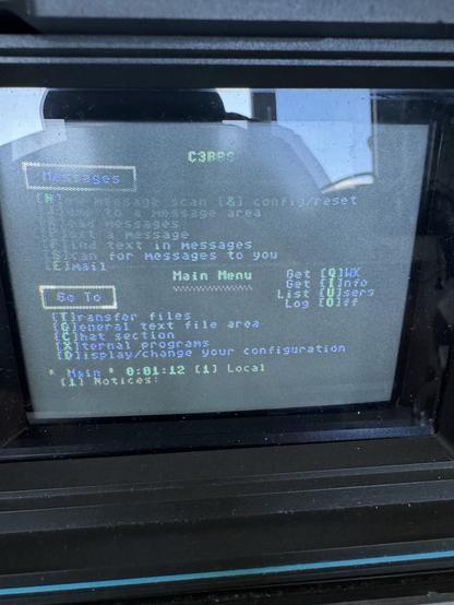 The image displays a retro computer screen with a text-based interface. It shows a "Main Menu" for a messaging system featuring options like viewing messages, sending a message, transferring files, and adjusting configurations. The background is dark with green text, typical