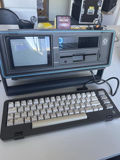 A vintage computer system featuring a compact design, a built-in display, and a keyboard. The screen displays text with a blue interface, while a disk drive is visible below the screen. A retro game cartridge is placed on top of the computer. The