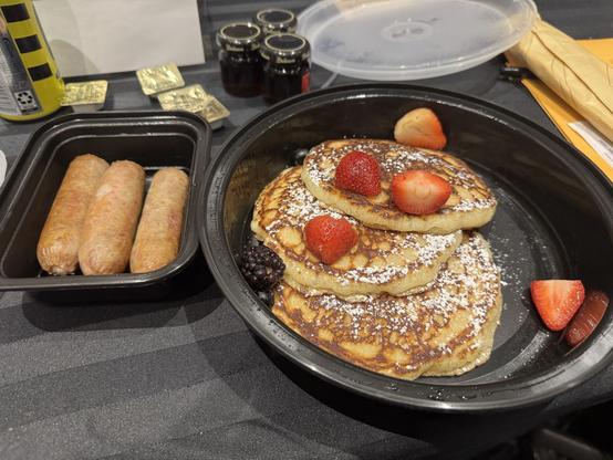 A to-go container with three large sausage links next to another with three substantial pancakes with strawberries on them. Behind them, 3 jars of maple syrup are visible