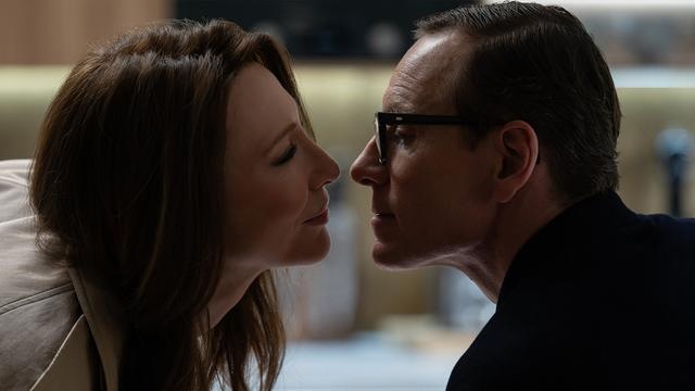 The image shows a close-up profile view of two people facing each other closely, appearing to be about to kiss. On the left, a person with long brown hair is smiling slightly. On the right, another person with short hair, wearing glasses, is also smiling subtly. The background is softly blurred, with neutral tones and soft lighting, creating a calm and intimate atmosphere.