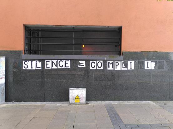 A collage graffiti saying "Silence = Complicity". A4 white paper, one capital letter painted in black per sheet. Pasted on an orange and grey wall. Some of the letters missing but still can read what it says.