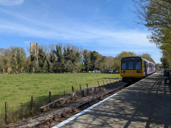 A BR Class 142 Pacer train sits in a country station platform. To the left is a grassy field with tall trees on the far side. The tall stone tower of a medieval abbey pines up above the trees.