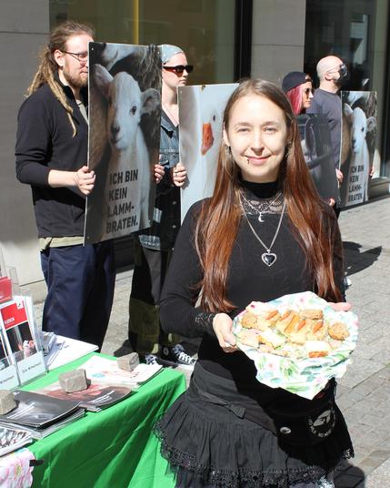 hinten stehen die Leute mit den Schildern, vorne ist ein veganer Infostand zu sehen, davor steht ein*e Aktivi mit einem Teller in der Hand auf dem verschiedene vegane Kuchenstücke und vegane Plätzchen liegen