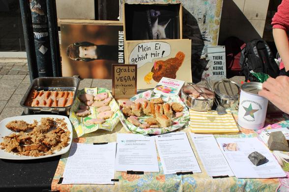 ein Foto vom Infostand, vegane Kuchenstücke mit einer kleinen Marzipanmöhre oben drauf liegen bereit, sowie vegane Kekse und die Rezepte dazu. Ein Aufsteller steht auf dem Tisch "gratis vegan Probieren". Hinten liegt ein Handgemaltes Plakat mit einem Huhn und dem Text: "Mein Ei gehört mir."