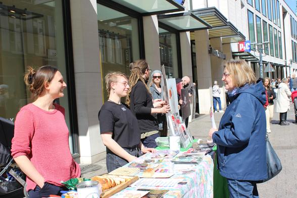 zwei Aktivis reden mit einer Frau am Infostand, die Sonne scheint und die Menschen wirken gut gelaunt