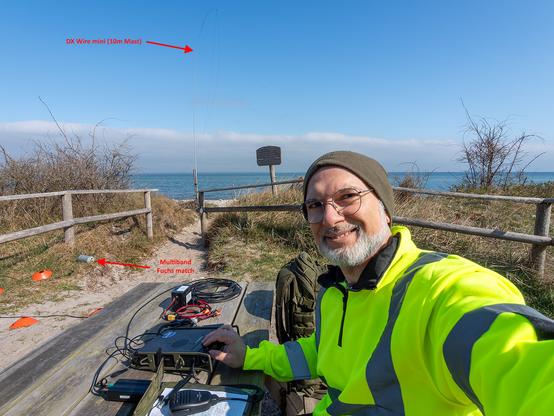 DJ4RAM sitting on a picnic table bench at the POTA spot near the dune beach at the baltic sea using HAM Radio equipment on the table: a Yaesu FT-857 with side rails, a CW keyer, a small LiFePO4 battery and coay cable. Ther are arrow marks on the ground pointig to a multiband fuchs match box and a 10m carbon mast holding the antenna wire as inverted V.