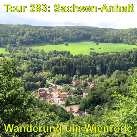 Blick von einem Aussichtspunkt auf ein Dorf in einem Tal, umgeben von dicht bewaldeten Hügeln. Die Dächer der Häuser sind vorwiegend rot, und die Gebäude gruppieren sich entlang einer Straße. Jenseits des Dorfes öffnet sich eine große Wiese auf den Hügeln. Das Grün der Bäume und Wiesen ist leuchtend und vielfältig. Das Licht ist klar.
