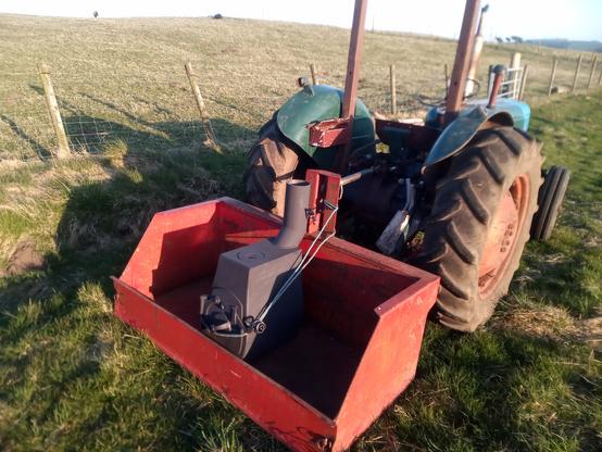The new stove arrives from the forge in the link box of The Little Tractor That Can. The tractor is standing on rough grass by a crooked fence and is lit with strong evening light.