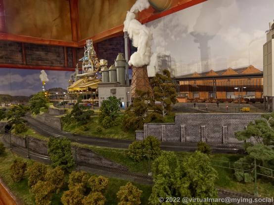 A photo of am odel railway. There are railway tracks in the front with trees. In the back is a steelworks with a smoking cooling tower.