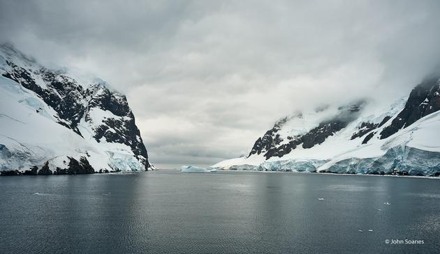 A muted palette with cloudy skies.  There are hues of blue in the white snow and the water is green-blue.  An ice floe can be seen floating in the middle.