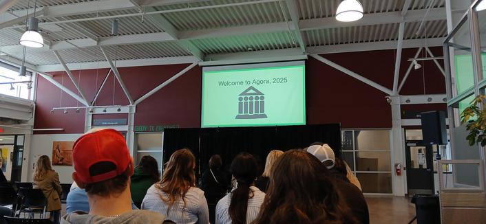 View from back row of a stage and display screen showing the Agora 2025 logo in the learning commons area at Red Deer Polytech