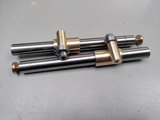 Two polished steel arms, one lays left to right, one the opposite. On one end of each is a small bronze bolt and about 12mm from the end is a slight groove, indicating a join.

Near the middle of each arm is a bronze bushing that sits round the arm. Perpendicular, and off centre a brass round piece sits. A thick steel washer and a stainless bolt are fitted to each of the brass parts.
