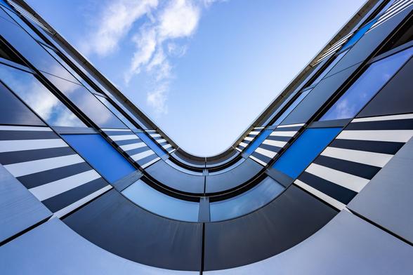Look-up symmetrical view towards the sky from a curvy corner of a modern building decorated with panels in black, blue or white or with black and white stripes, in the Beaugrenelle quarter in Paris. The windows of the building reflect the blue skye and a few small white clouds.
