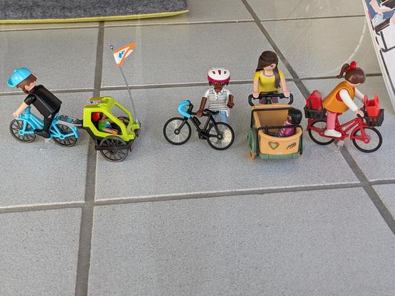 Vier Playmobil Figuren nebeneinander. Von links nach rechts.
Eine Figur auf einem Fahrrad mit Anhänger. Eine Figur steht und hat ein Fahrrad in der Hand. Eine Figur sitzt auf einem Lasten Dreirad mit Korb in der Front, darin sitzt eine kleine Figur. Eine Figur sitzt auf einem Fahrrad das vorne und hinten jeweils einen Korb hat.