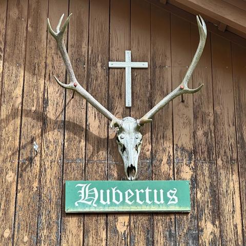 A cross hangs above an antlered deer skull which in turn hangs above a green sign that reads “hubertus” in a blackletter font on the weathered wood exterior wall of a small hut in central Switzerland.