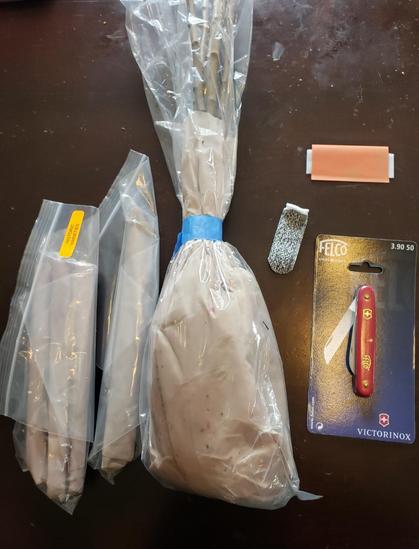Assorted items needed for grafting placed on top of a wooden table. Items are lined up and include two ziplock baggies of scion wood tightly wrapped in paper, one large bag  with the rootstock, thumb protector, grafting tape and knife.