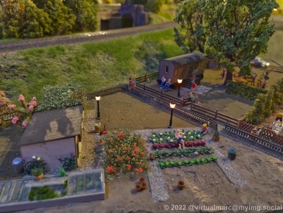 A photo of a garden area of a model railway with sheds, greenhouses and flowerbeds. Multiple people caring about thos gardens.
