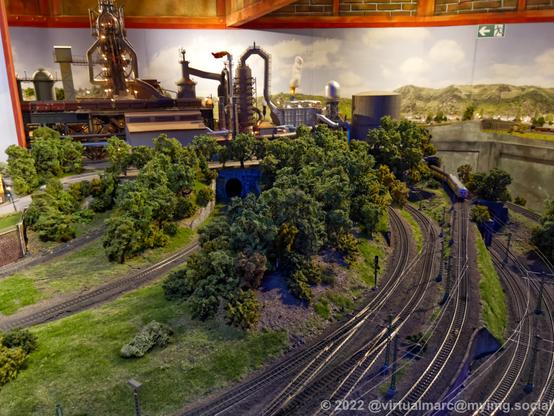 A photo of the back of a steelworks of a model railway with multiple train tracks and a passenger train driving away from the camera.