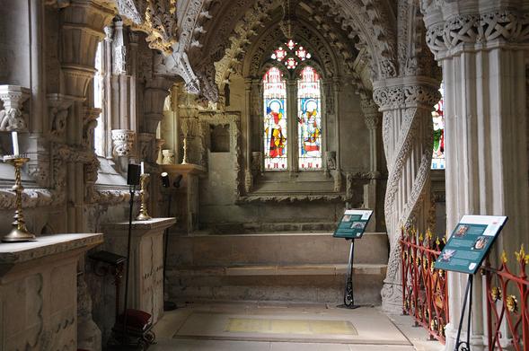 Part of the interior of Rosslyn Chapel. The image shows the interior of the end of the chapel with windows on the left and two columns on the right. Another window is directly ahead. There are interpretive panels on stands on the right. Much of the stonework is intricately decorated.