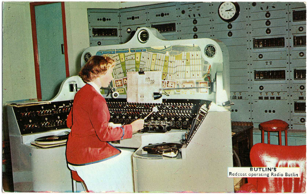 A  woman in a coat the colour of tomato ketchup, sitting in front of an array of electro-mechanical switches, controlling a map of a holiday camp. There is a mini xylophone and a sleek phone probably stolen from the set of UFO