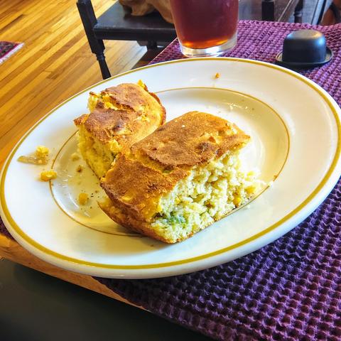 A yellow-rimmed ceramic plate holds 2 pieces of cornbread, one of which is missing a bite.  The sliced edges of the cornbread show that it has green onion & corn kernels inside.  

The plate is lying on a maroon waffle-weave cloth napkin, which itself is on a wooden TV table.  Immediately behind the plate, one can see part of a glass of iced tea, & a miniature black plastic bowlers hat.
